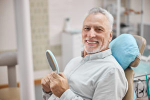 Cheerful patient sitting in a dental chair and holding a mirror while smiling at the camera